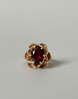 A gold ring with a textured, flower-like setting holding a faceted dark red gemstone, resting on a wooden surface with a gray background.