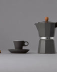 Black stovetop espresso maker with wooden handle and matching cup on a gray background