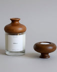 A clear glass candle jar with a rounded brown wooden lid and a white label reading 'KÖKLI', shown beside a matching brown wooden candle holder on a dark surface against a light gray textured wall.