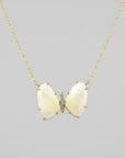 A delicate gold chain necklace featuring two pale yellow-green butterfly wing charms connected by a small gold heart, displayed on a textured blue denim background.