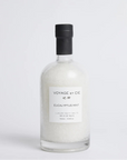 A clear glass bottle filled with white bath salts, featuring a minimalist white label and a black cap, set against a seamless light gray background.