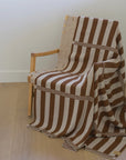 A woven throw blanket with brown and cream vertical stripes draped over a wooden armchair in a bright room with light wood floors and a window showing green foliage outside.