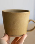 A hand holding a matte ceramic mug with a warm sandy beige finish and a matching loop handle, set against a soft, out-of-focus light background.