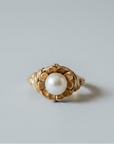A vintage gold ring featuring a central round white pearl surrounded by a floral petal setting, resting on a dark gray surface.