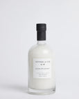 A clear glass bottle filled with white bath salts, featuring a minimalist white label and a black cap, set against a seamless light gray background.