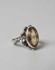 A silver-toned ring with an ornate floral band and a large oval faceted amber gemstone, resting on a brown wooden surface against a textured off-white wall.