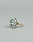 A gold halo ring featuring a round light blue gemstone surrounded by small clear stones, resting on a brown wooden surface with a dark gray background.