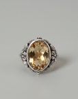 A vintage-style silver ring with an oval faceted yellow gemstone, resting on a brown wooden surface against a textured off-white wall.