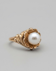 A vintage-style gold ring featuring a large, round, lustrous white pearl set in an ornate, textured gold bezel, resting on a dark gray surface.